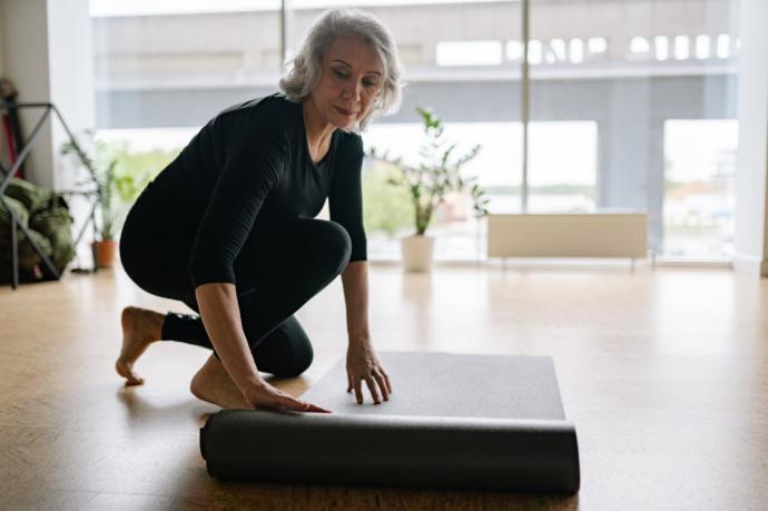 Femme senior cheveux blancs et tenue noire agenouillée qui déroule un tapis de yoga noir 
