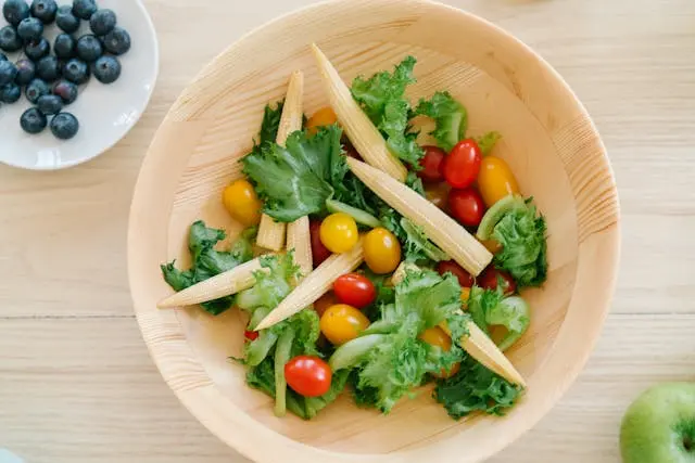 Vue de haut saladier en bois avec légumes et coupelle de myrtilles et une pomme verte à côté, sur table en bois clair