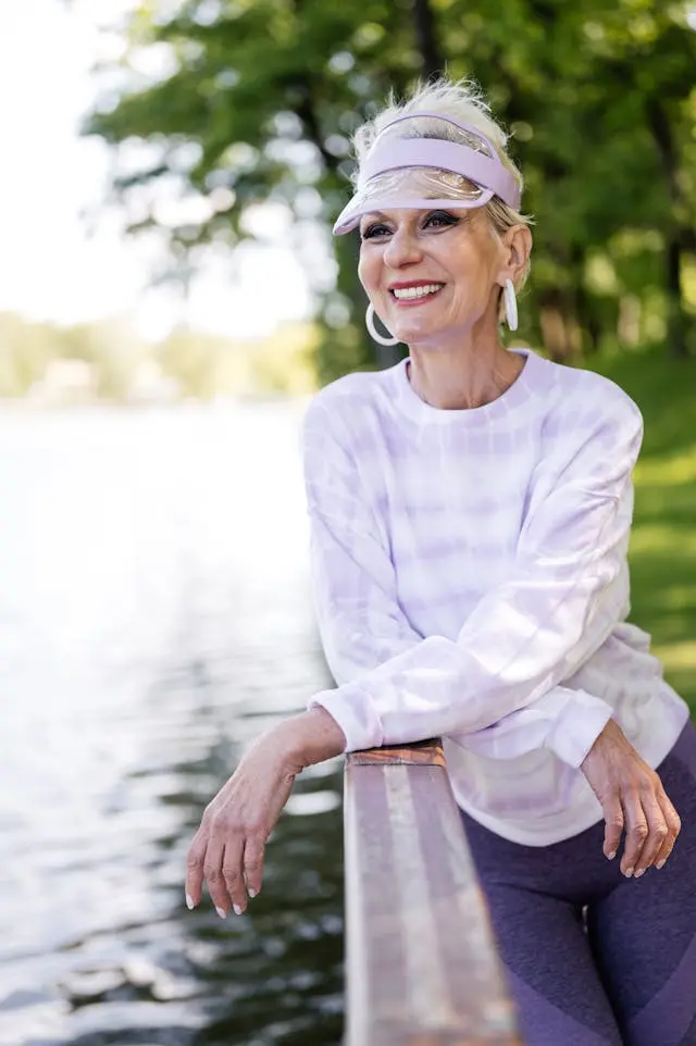 Femme senior en tenue de sport violette et mauve avec casquette à visière et boucles d'oreille créoles blanches qui sourit accoudée à une balustrade avec de l'eau et des arbres en arrière-plan